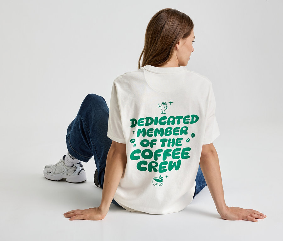Žena sedí na zemi, má na sobě tričko s nápisem „Dedicated Member of the Coffee Crew“, džíny a tenisky.