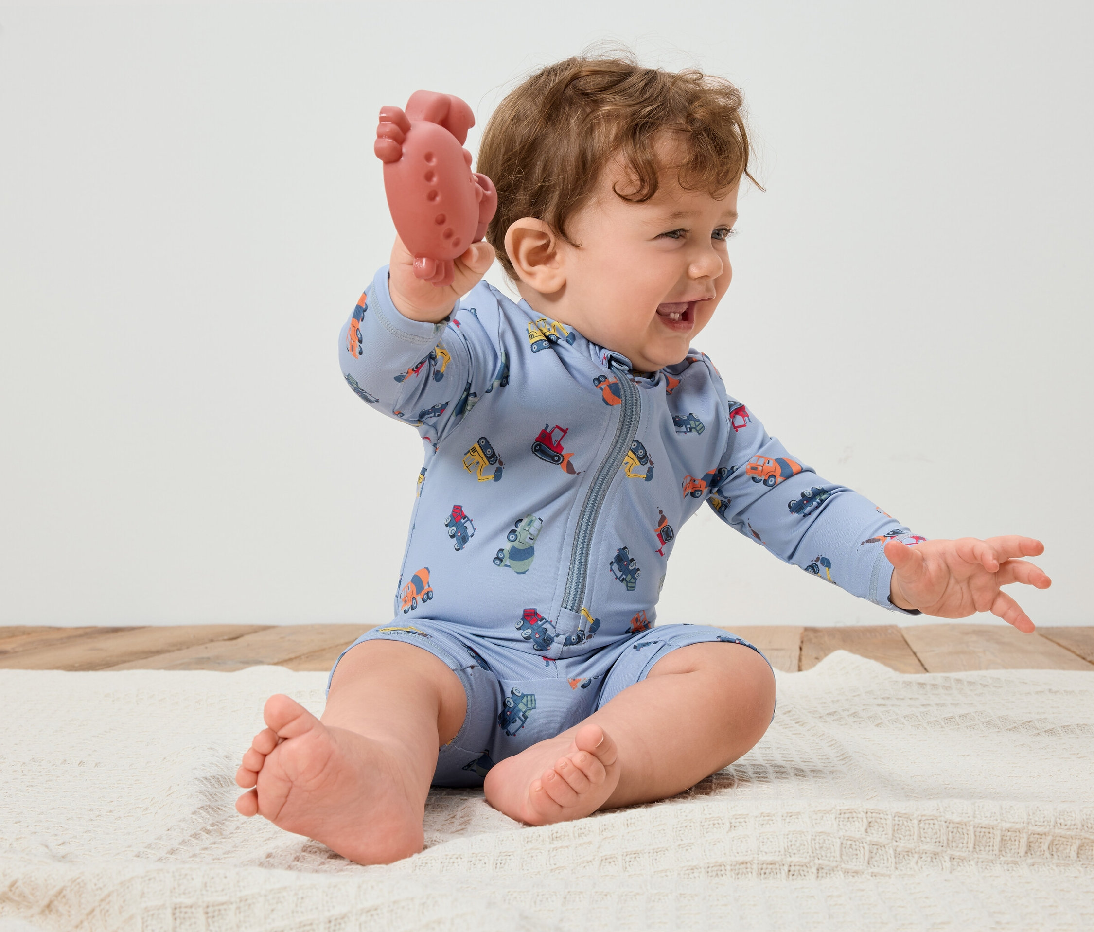 Usmívající se kojenec s kudrnatými vlasy sedí na bílé dece a v ruce drží červené kousátko. Kojenec má na sobě světle modrý overal s barevnými vozidly.