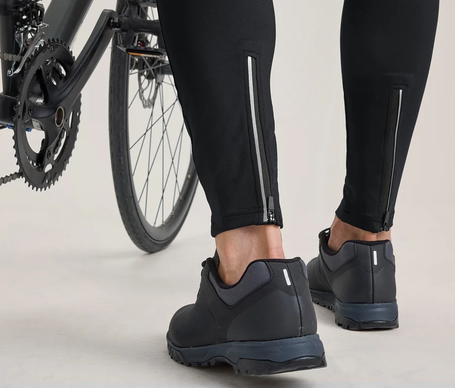 Spodní části nohou a chodidla osoby stojící vedle jízdního kola. Osoba má na sobě černé kalhoty s reflexními zipy u kotníků a černé cyklistické boty.