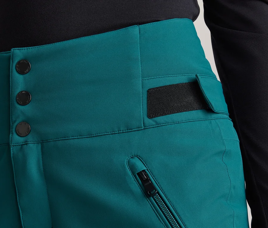 Detail osoby v tyrkysových kalhotách s vysokým pasem a černém topu. Kalhoty mají tři černé knoflíky, kapsu na zip a klopu se suchým zipem.
