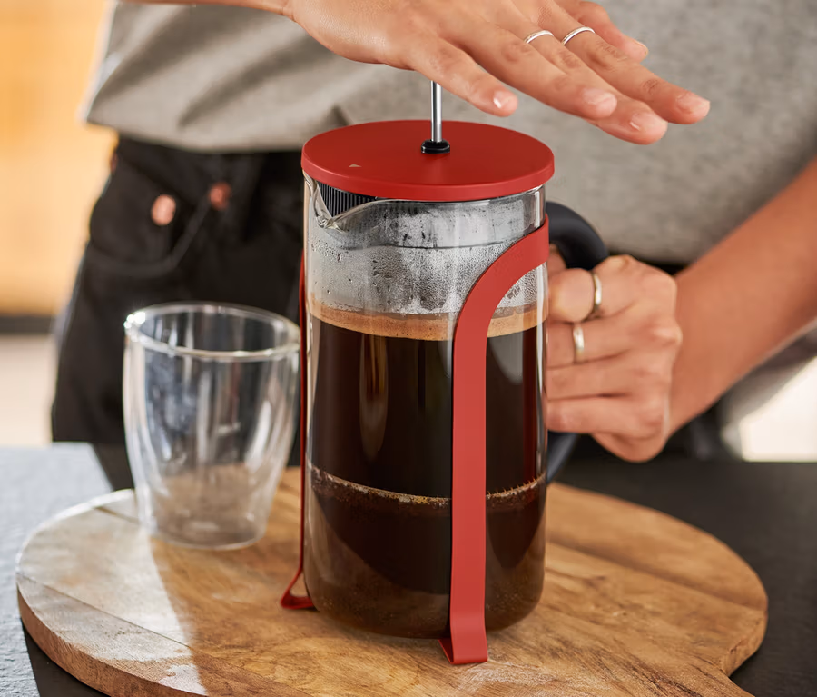 Žena stlačuje červenou konvičku french press 800 ml na 6 šálků na dřevěném prkénku.