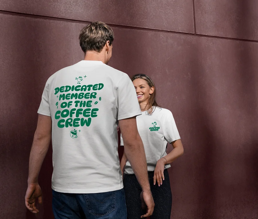 Muž v bílém Unisex tričku Coffee Crew se zeleným nápisem "DEDICATED MEMBER OF THE COFFEE CREW" na zádech. Žena v pozadí se usmívá a má také bílé tričko s logem "Coffee Crew".