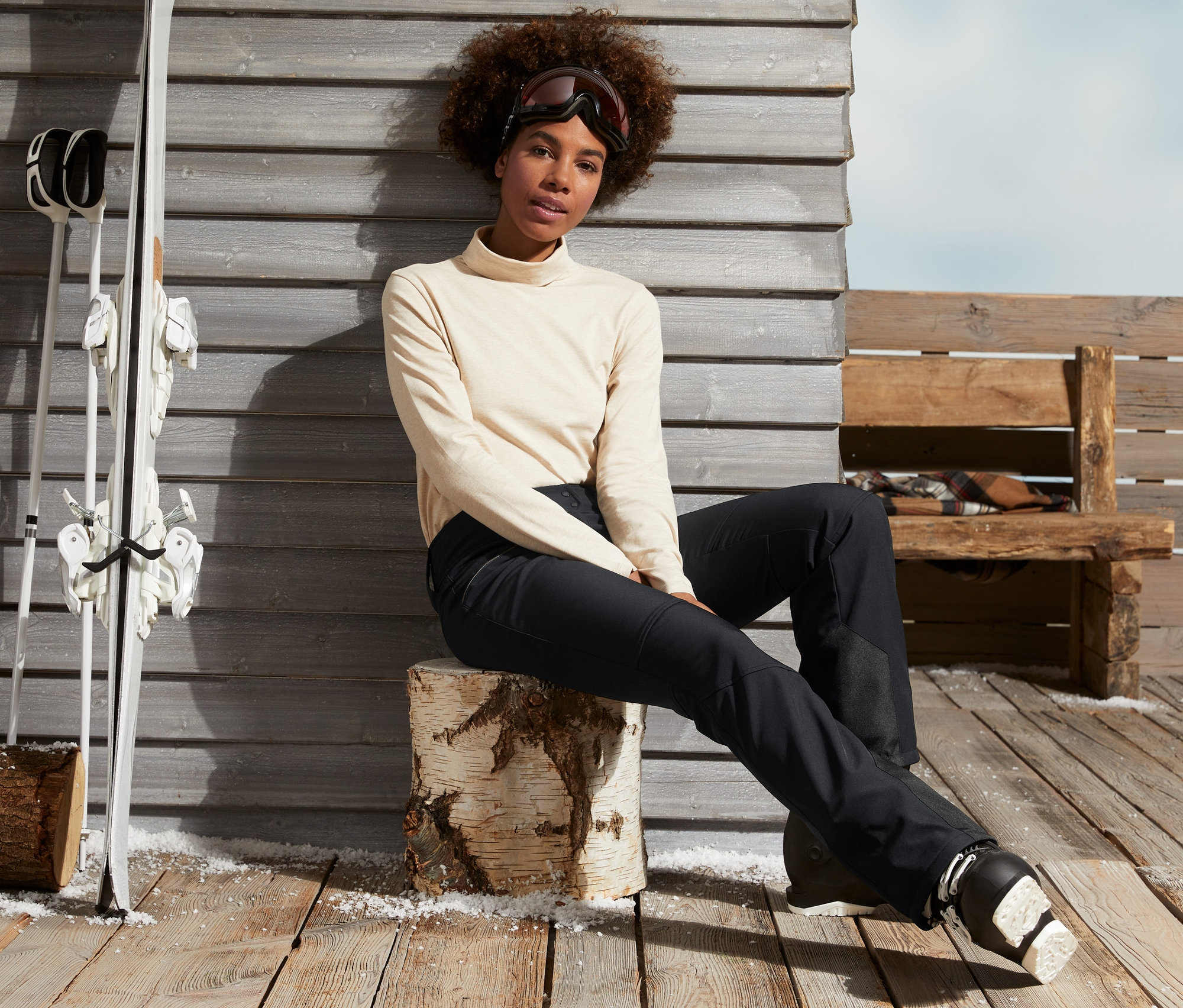 Žena sedí na dřevěném špalku v černých lyžařských kalhotách a lyžařských botách.