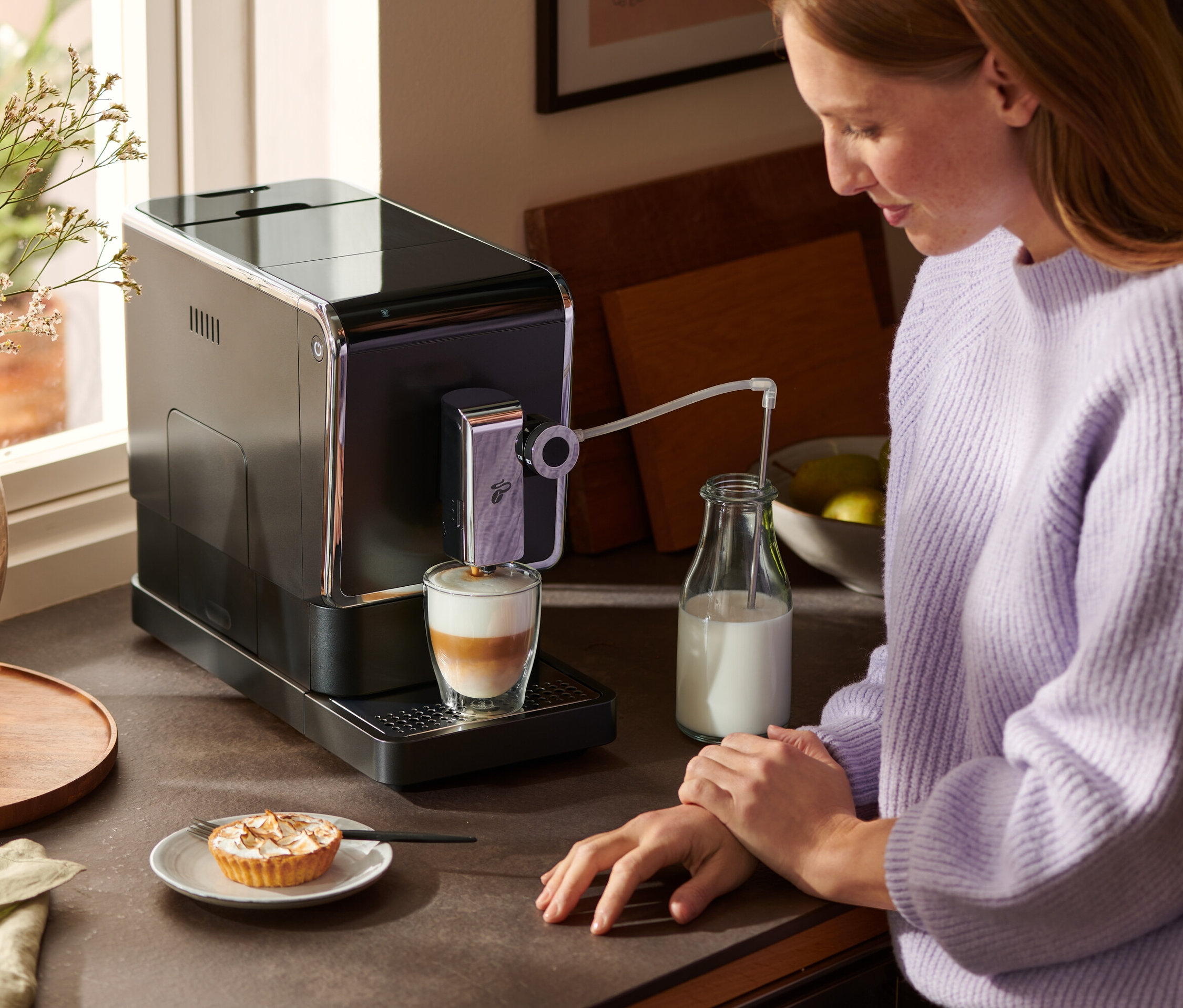 Žena stojí u linky a používá antracitový kávovar Tchibo »Esperto Pro« k přípravě latté. Vedle kávovaru stojí láhev mléka a na talíři je koláč.