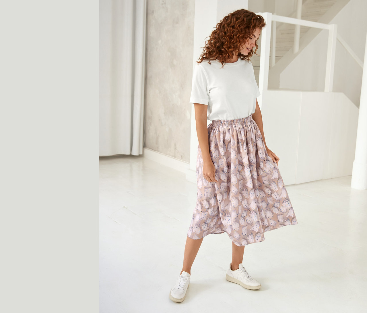 Žena stojí v bílé místnosti a má na sobě bílé tričko NAH/STUDIO s potiskem z biobavlny a vzorovanou sukni.