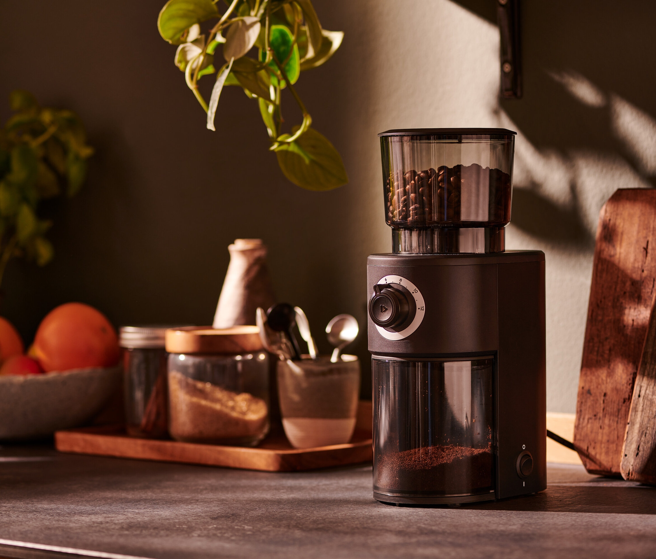 Hnědý elektrický mlýnek na kávu stojí na kuchyňské lince vedle sklenic s kořením.
