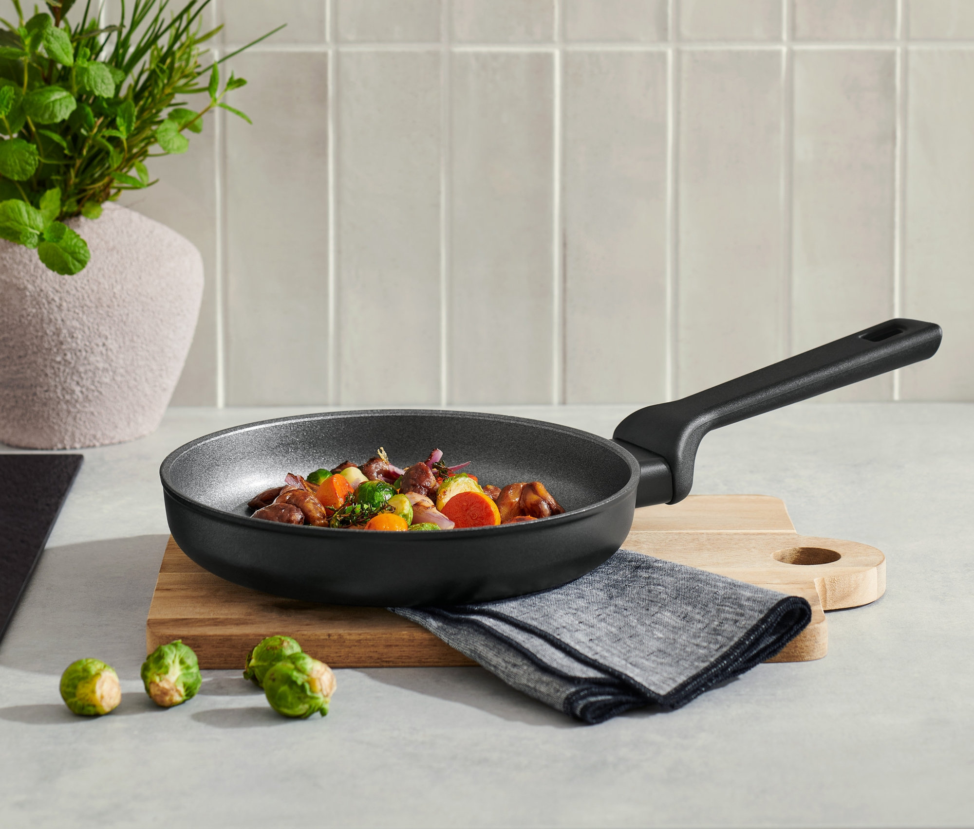 Na kuchyňské lince stojí profesionální hliníková pánev o průměru cca 20 cm s obsahem, vedle ní leží tři růžičkové kapusty.