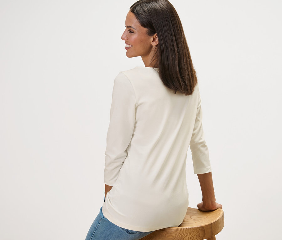 Žena sedí bokem na dřevěné stoličce a má na sobě bílé tričko s tříčtvrtečním rukávem.