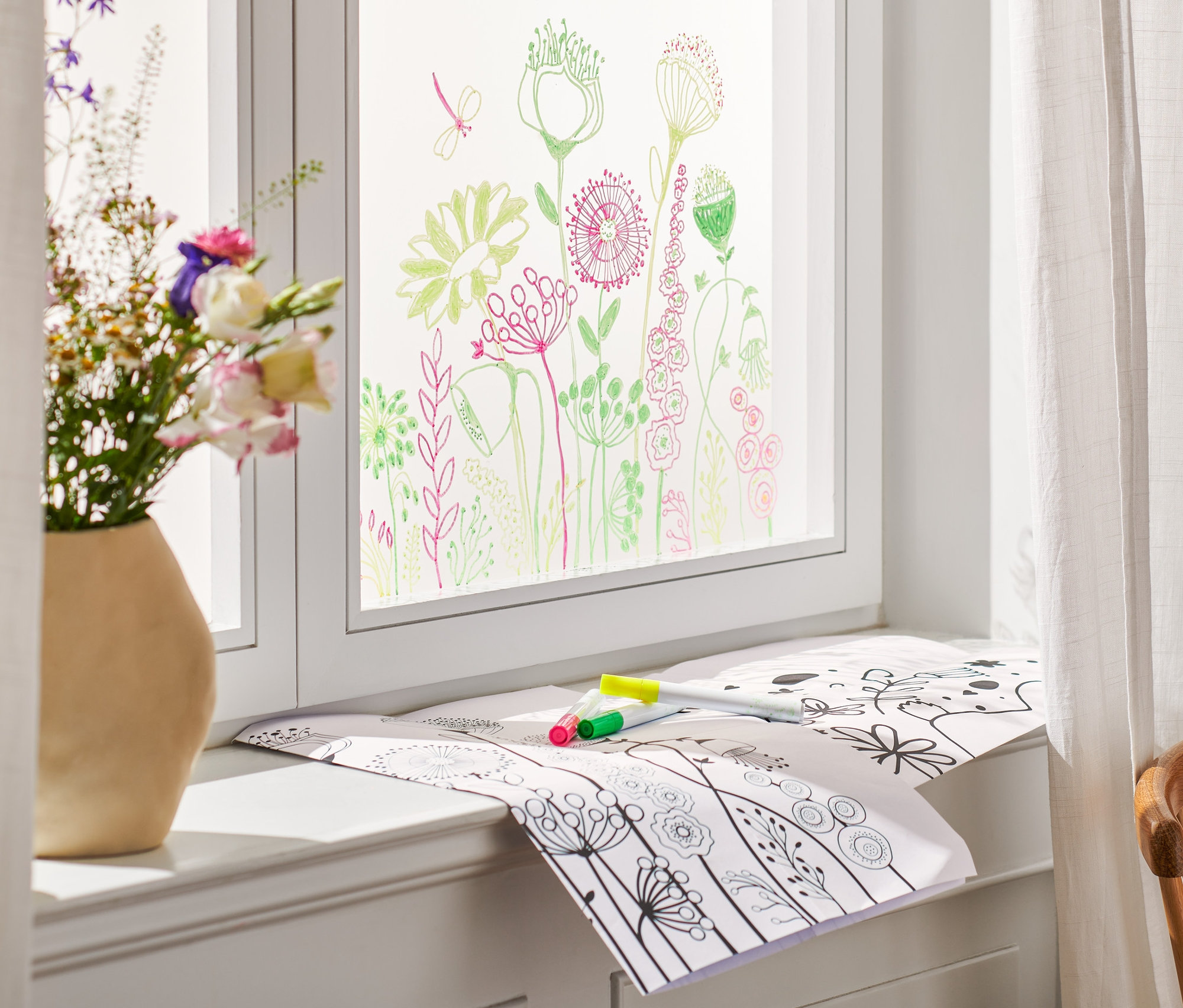 Květiny ve váze stojí na okenním parapetu, okno je pomalované a na parapetu leží sada na velikonoční kresby do oken a fixy.