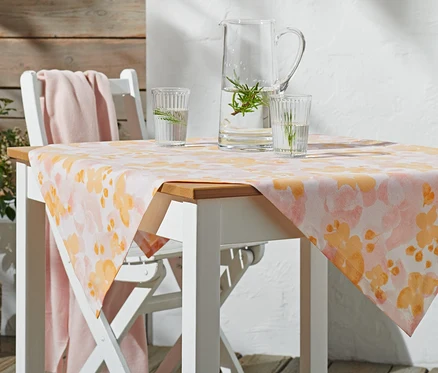 Na bílém zahradním stole leží středový ubrus cca 90 x 90 cm s oranžovým květinovým vzorem. Na stole stojí skleněný džbán a dvě sklenice s vodou.
