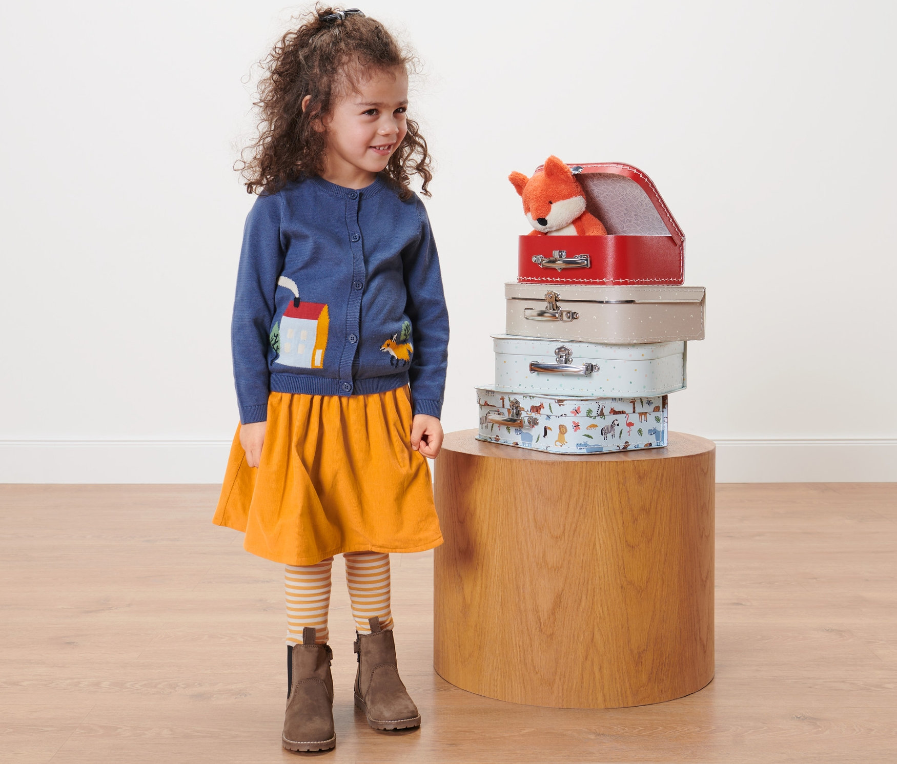 Dívka stojí vedle stohu kufříků, má na sobě dětskou oboustrannou manšestrovou sukni.