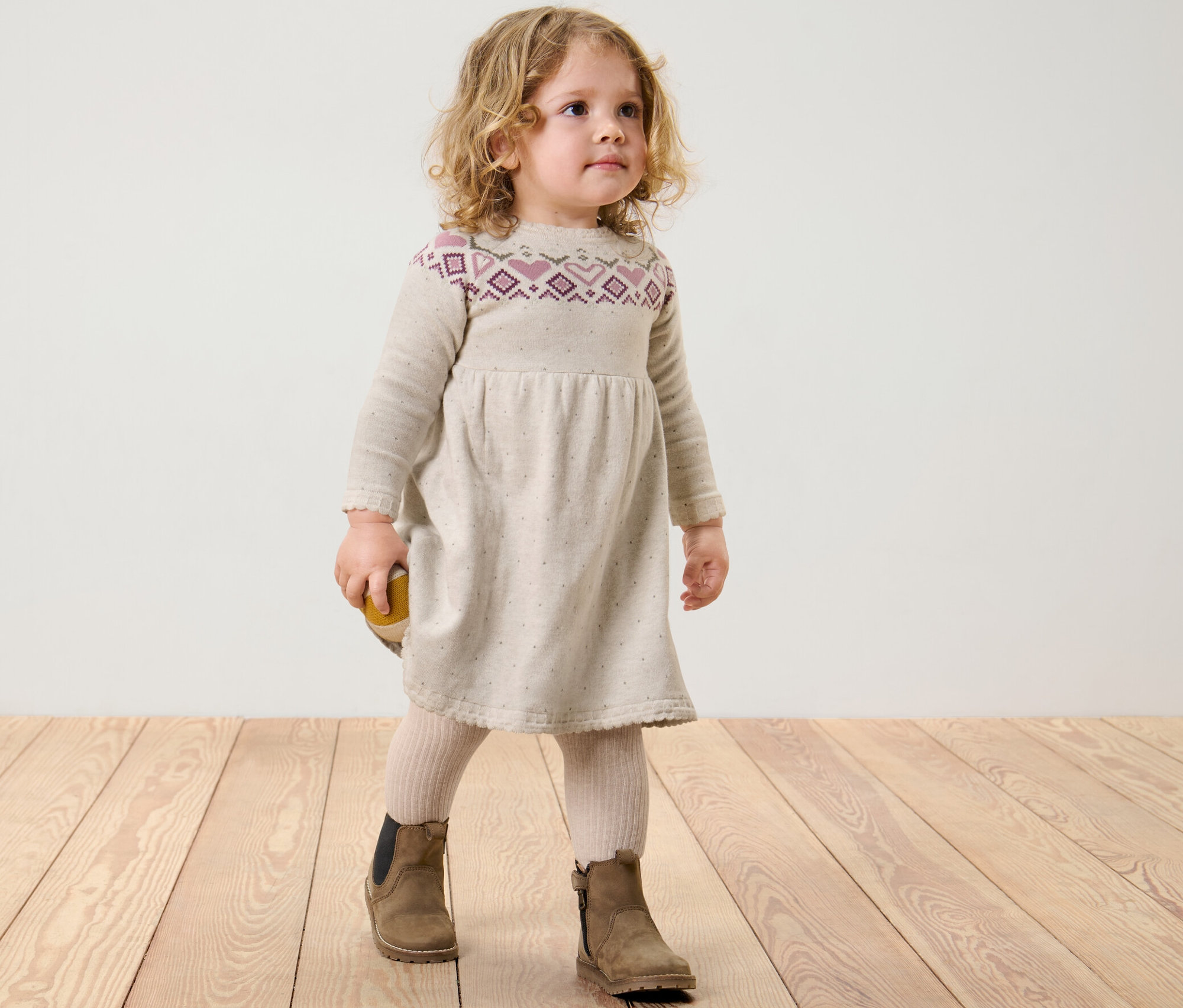 Dívka stojí na dřevěné podlaze a má na sobě pletené šaty.