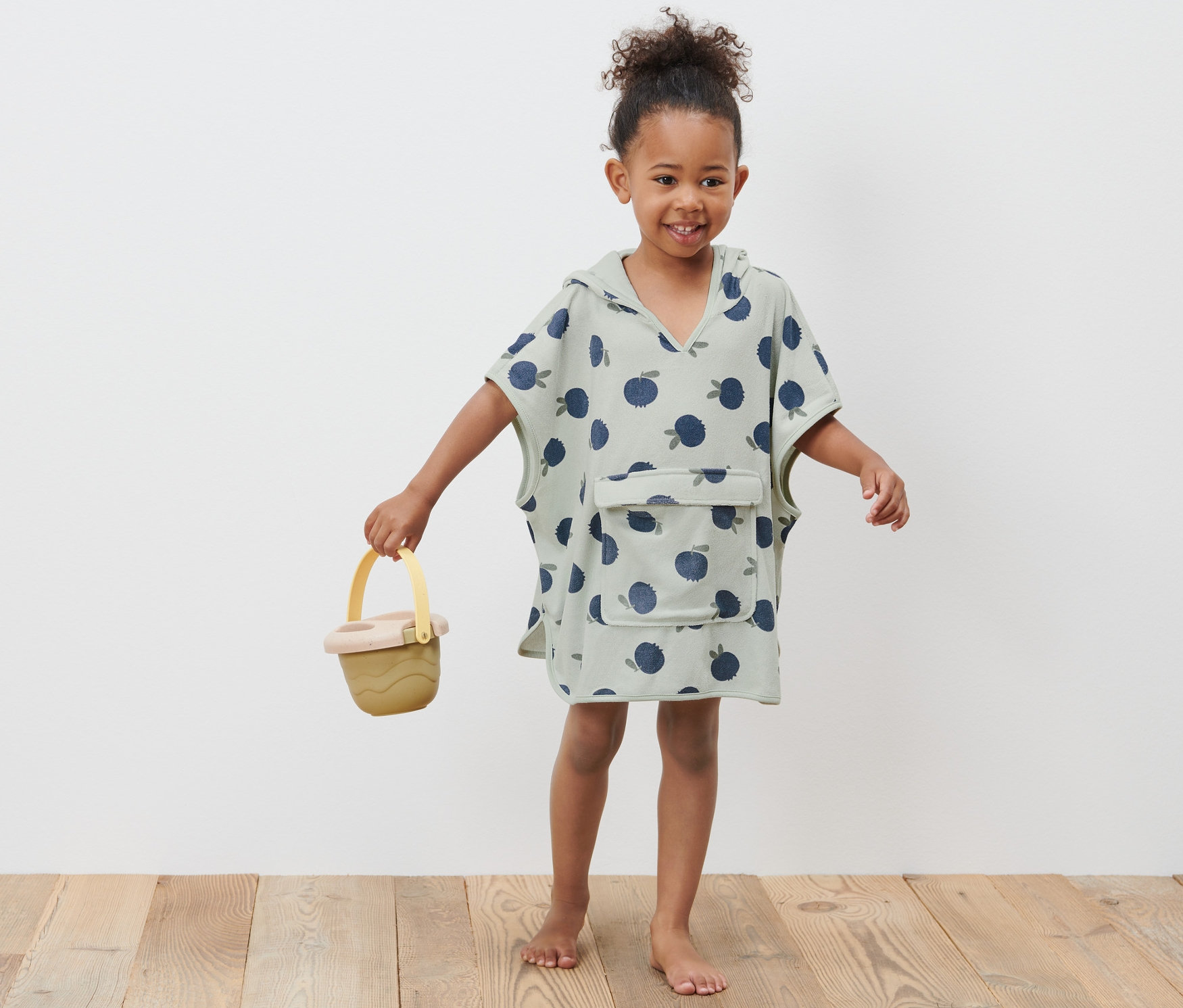 Usmívající se holčička v dětském froté ponču drží žlutý kyblík.