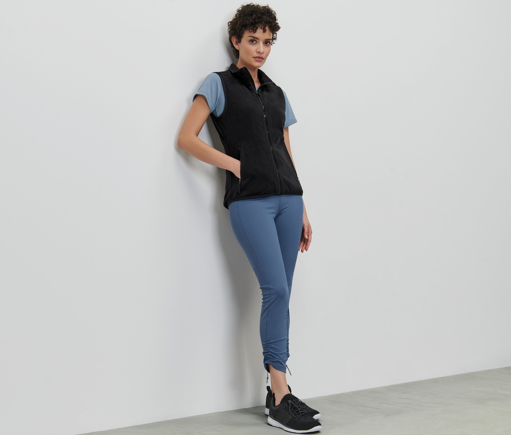 Žena opřená o stěnu má na sobě fleecovou vestu, tričko a legíny.