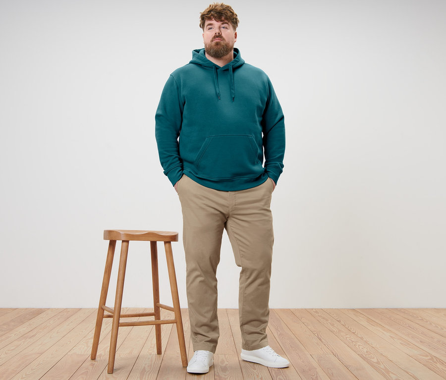 Muž stojí a má ruce v kapsách. Má na sobě petrolejovou mikinu s kapucí a béžové chino kalhoty.