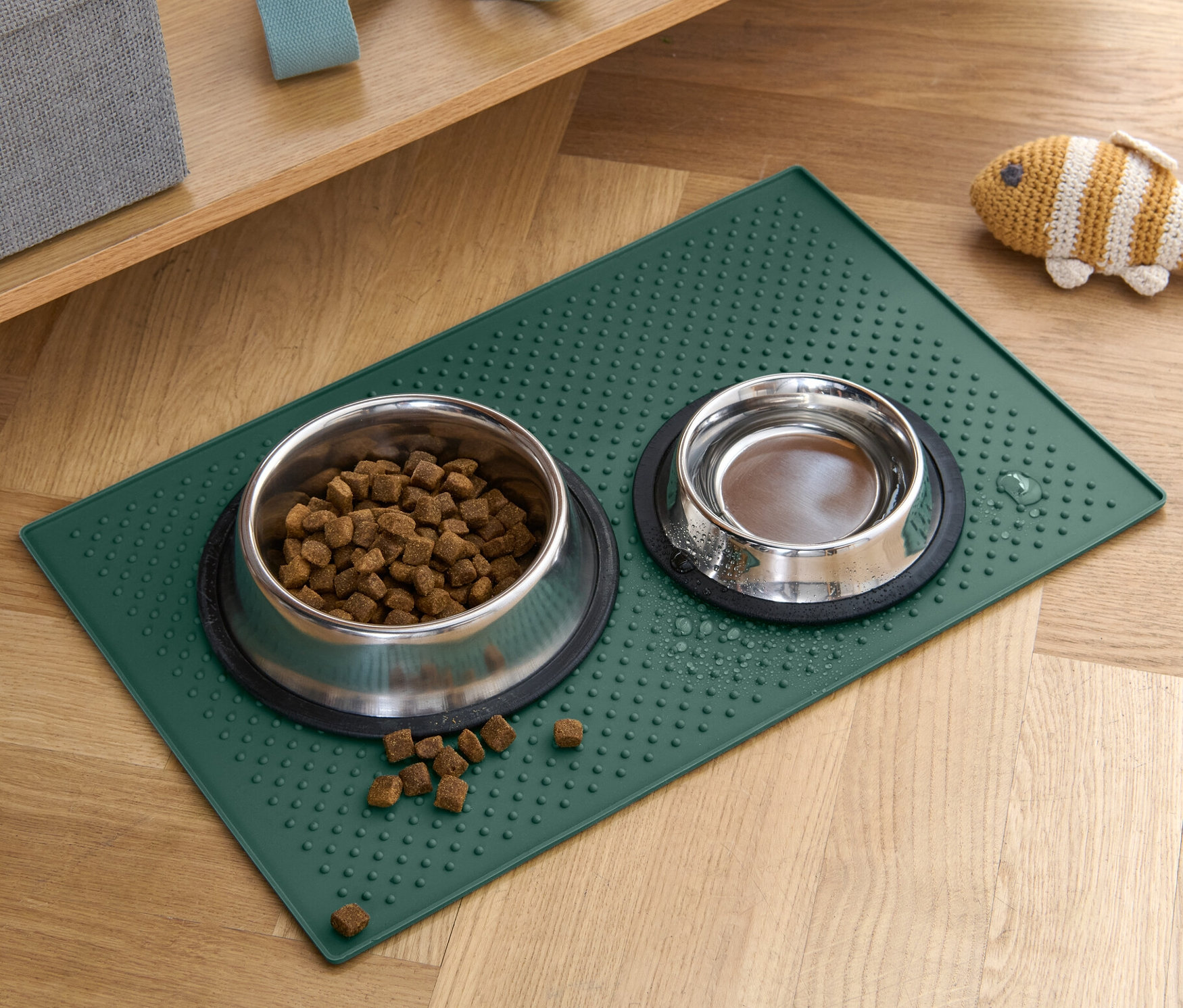 Dvě misky pro zvířata stojí na zelené podložce pod misku na krmivo na dřevěné podlaze.