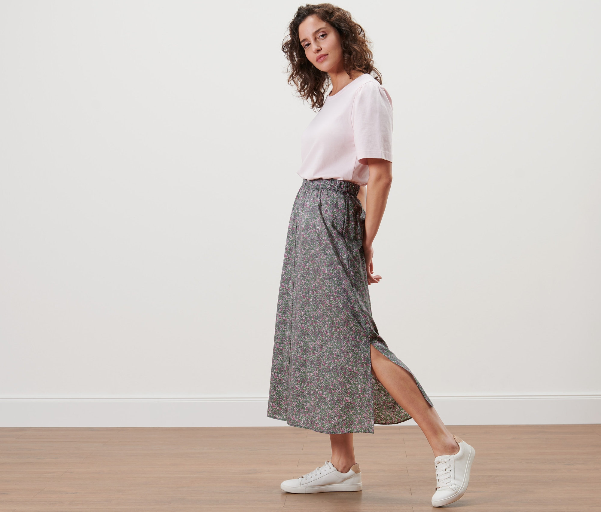 Žena pózuje v květované sukni midi a bílých teniskách.