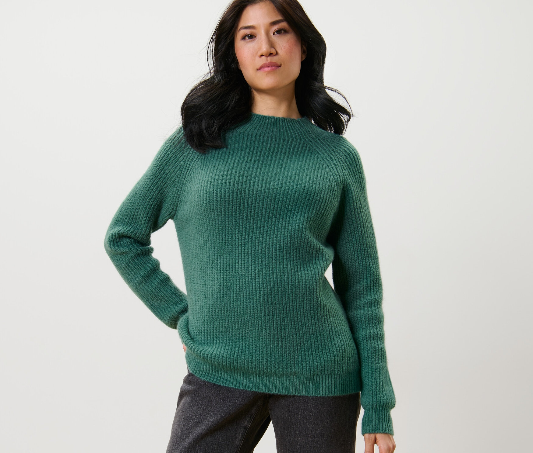 Žena pózuje v zeleném svetru z hrubého úpletu se stojáčkem a tmavých kalhotách.