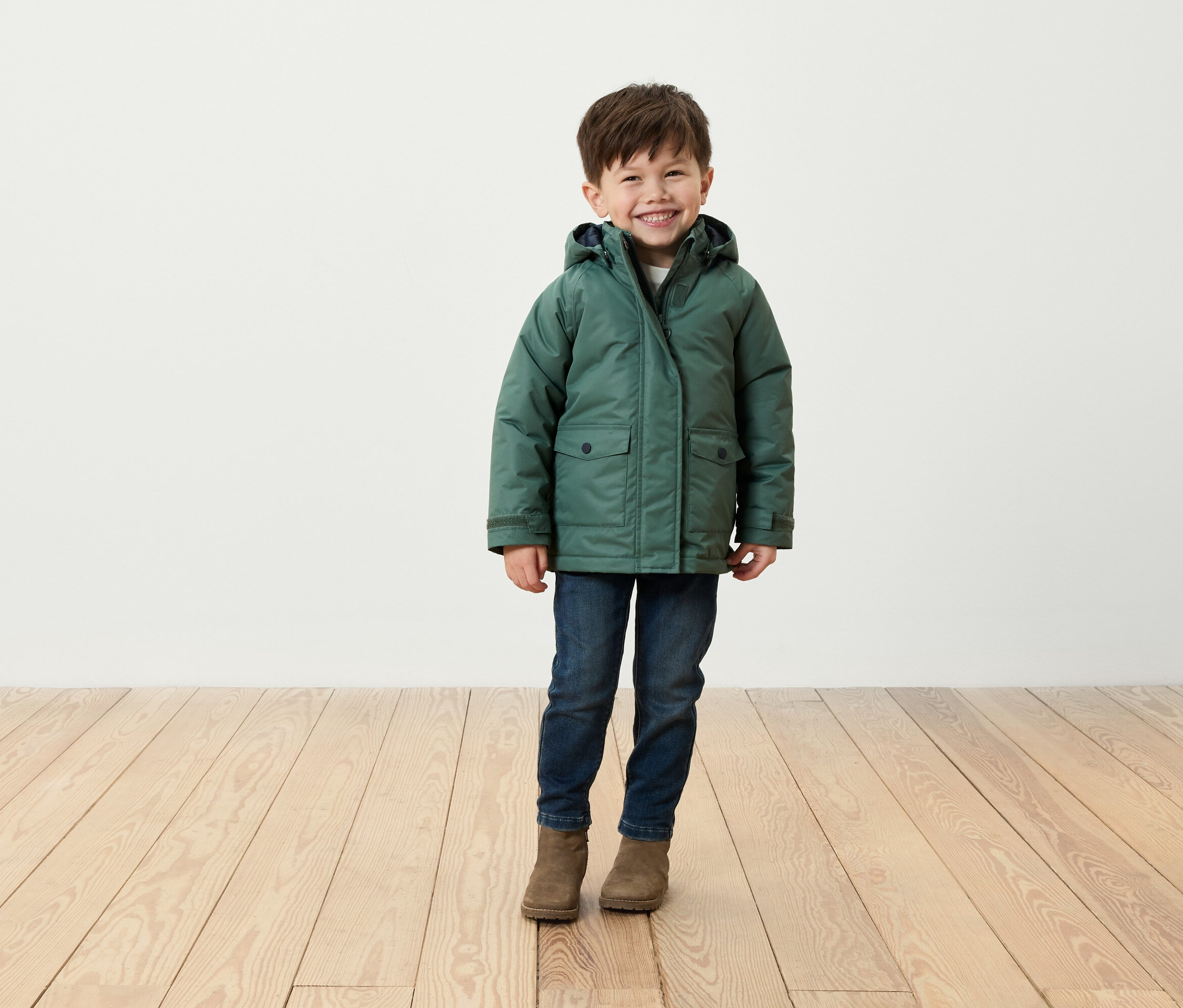 Usmívající se chlapec stojí na dřevěné podlaze a má na sobě zelenou bundu, džíny a hnědé boty.