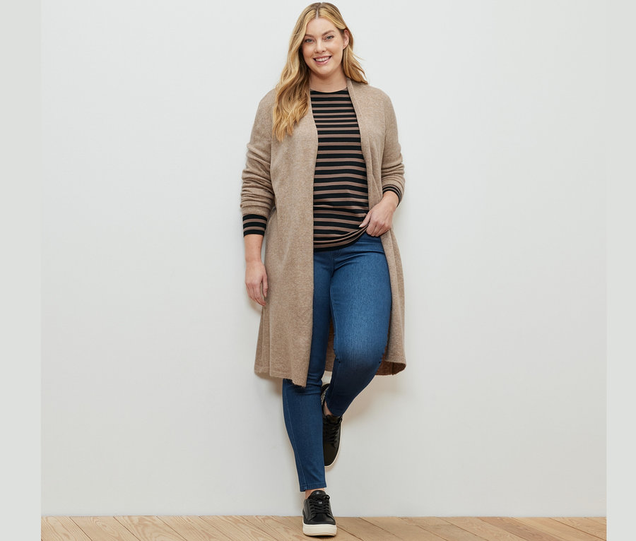 Žena pózuje v džegínách, tričku s dlouhým rukávem s černými a hnědými proužky a otevřeném dlouhém kardiganu.