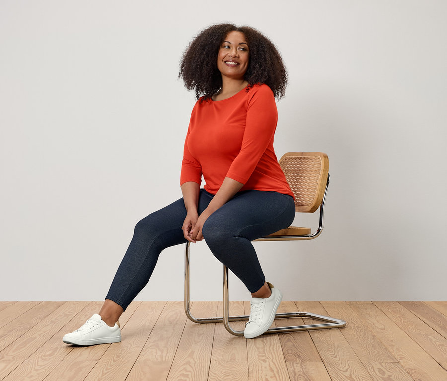 Žena sedí na židli, má na sobě oranžovou halenku, tmavě modré legíny a bílé tenisky.