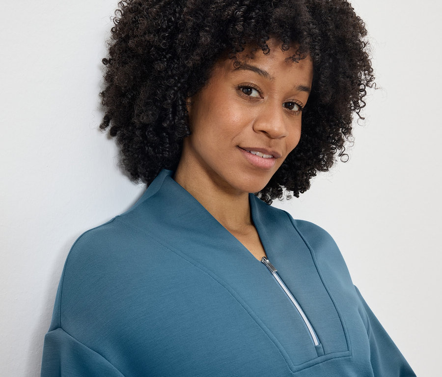 Portrét usměvavé ženy s afro účesem v modrozelené mikině se zipem.