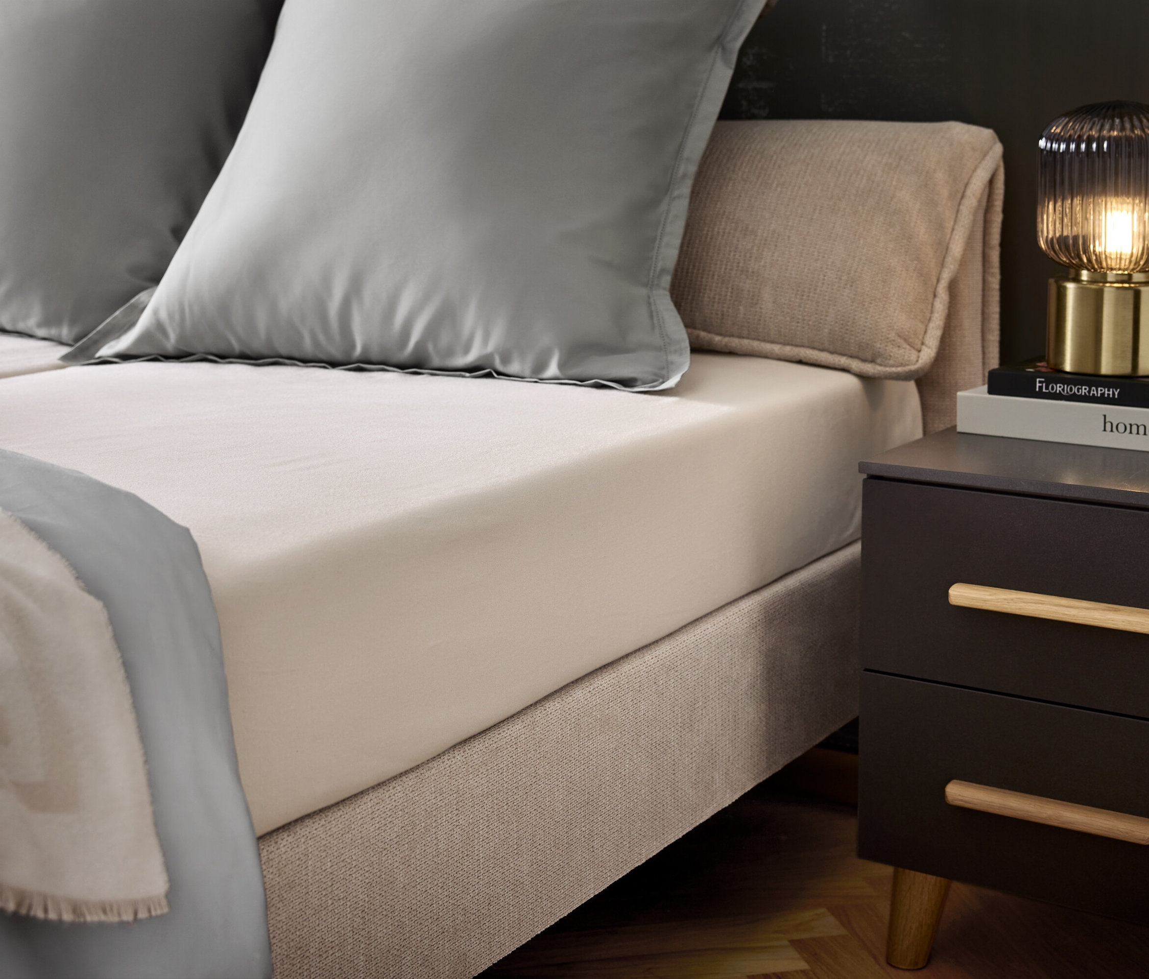 Ložnice s postelí, dvěma šedými polštáři a šedým přehozem. Na nočním stolku lampa a knihy.