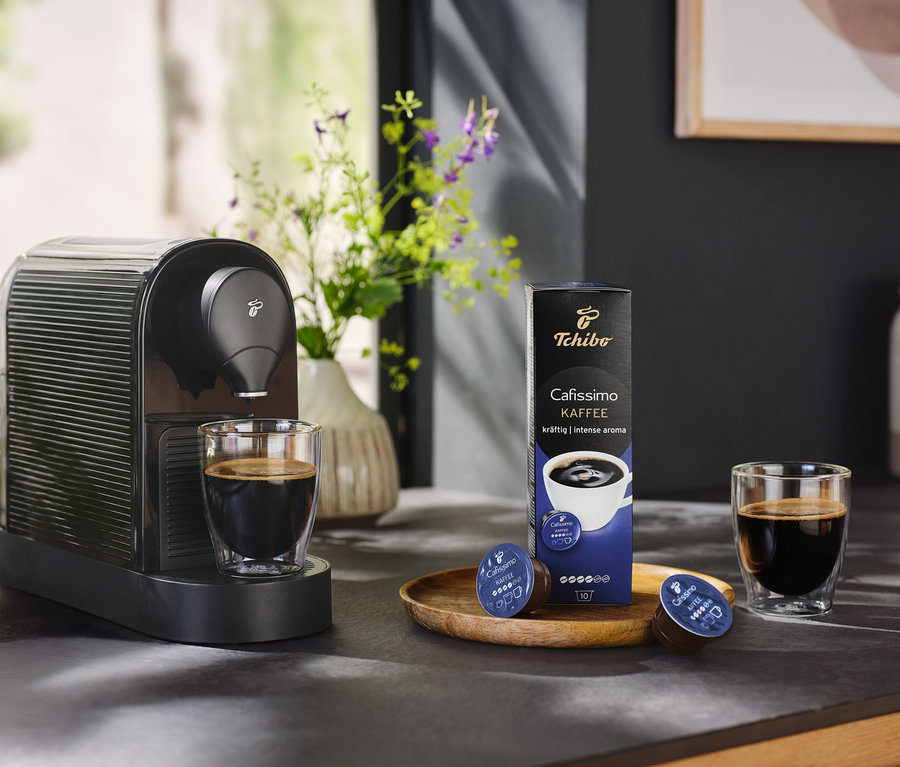 Černý kávovar Cafissimo pure+ s šálkem kávy a balením kávových kapslí.