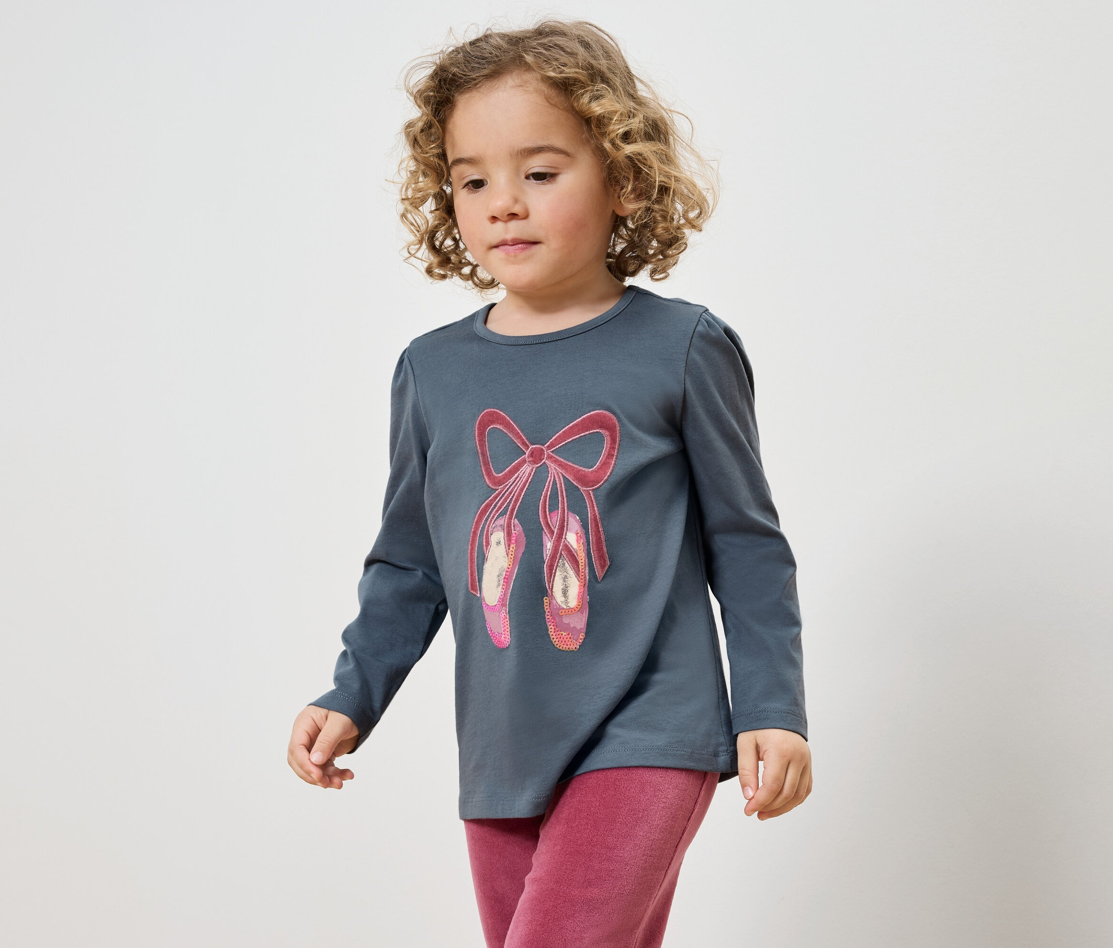Holčička s kudrnatými vlasy v šedém tričku s obrázkem baletek a růžových kalhotách.
