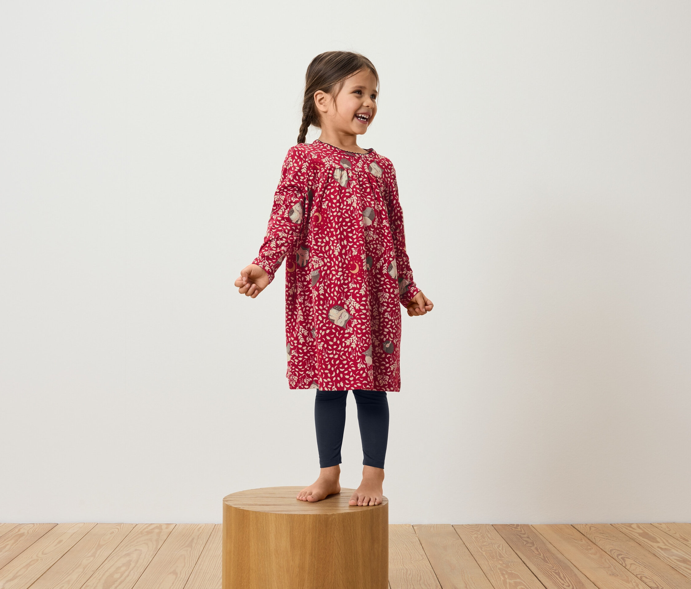 Usmívající se dívka stojí na dřevěném podstavci a má na sobě červené šaty s dlouhým rukávem a tmavě modré legíny.