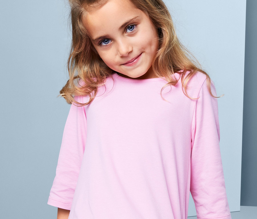 Dívka s hnědými vlasy a modrýma očima nosí růžové šaty. Ve vlasech má gumičku do vlasů.