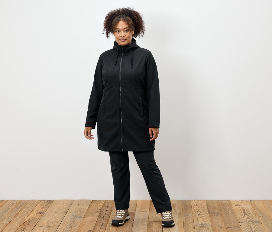 Žena v černém kabátě a kalhotách stojí na dřevěné podlaze.