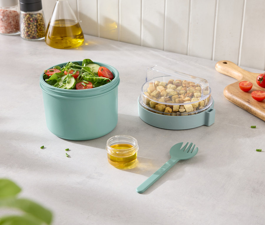 Zeleninový salát v tyrkysové nádobě s krutony v nádobě s průhledným víkem a dresinkem a vidličkou vedle.