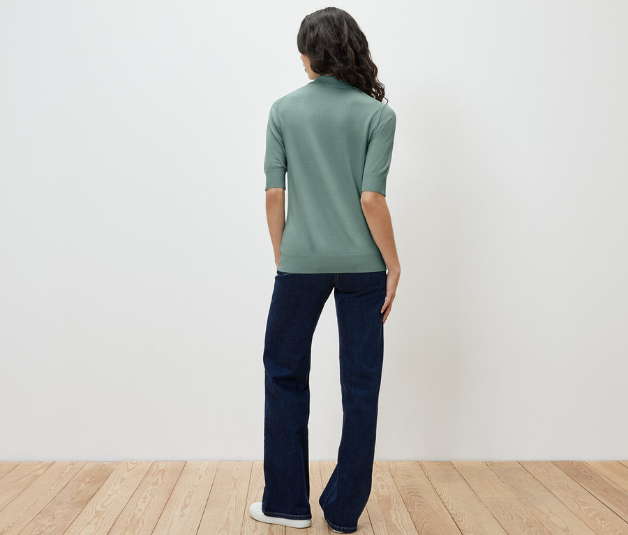 Žena stojí zády k fotoaparátu a má na sobě světle zelenou polokošili z jemného úpletu a džíny.