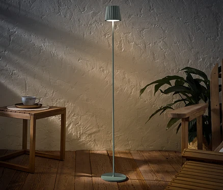 Šalvějová LED dobíjecí venkovní stojací lampa stojí vedle dřevěného stolku s šálkem a dřevěného křesla.