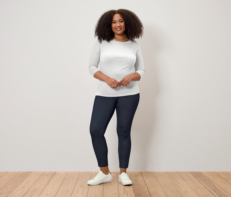 Žena s kudrnatými vlasy stojí na dřevěné podlaze a má na sobě bílé tričko s tříčtvrtečními rukávy a tmavě modré legíny.