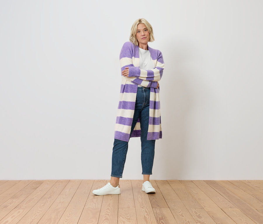 Blonďatá žena stojí a má zkřížené ruce. Na sobě má džíny střihu "Nyla" středně modré barvy a pruhovaný fialovo-bílý kardigan.