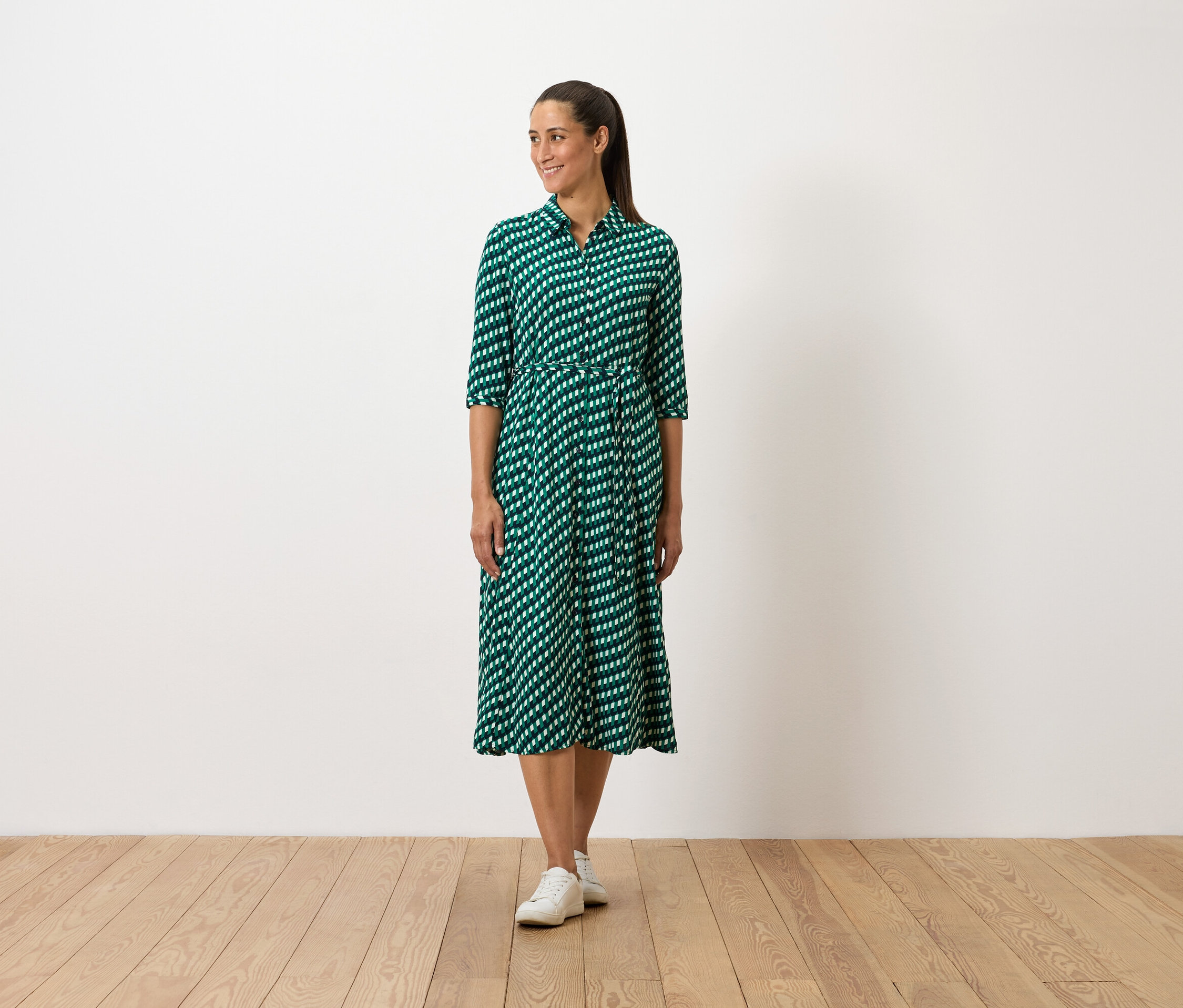 Žena stojí a má na sobě zelené šaty se vzorem a bílé tenisky.