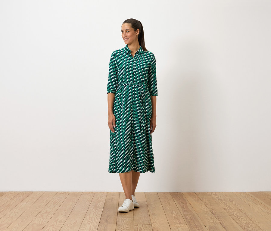 Žena stojí a má na sobě zelené šaty se vzorem a bílé tenisky.