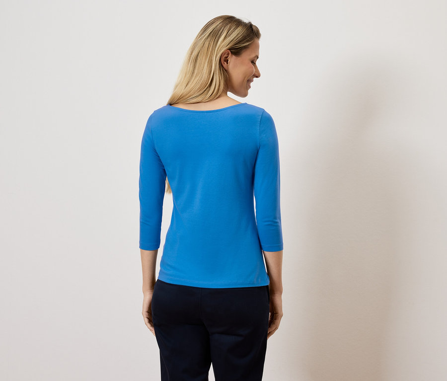 Žena se dívá přes rameno a má na sobě modrý top s tříčtvrtečním rukávem a tmavé kalhoty.