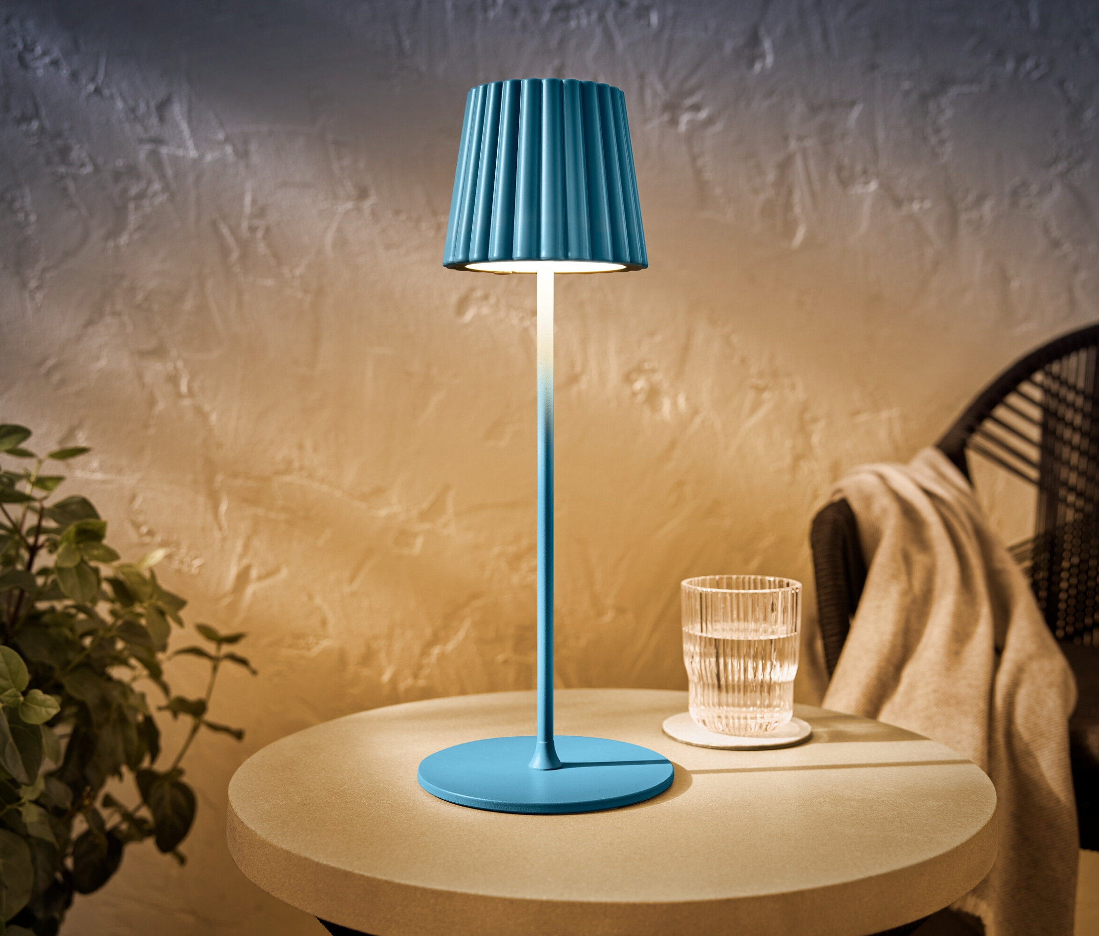 Modrá LED venkovní dobíjecí stolní lampa stojí na stole vedle sklenice vody.