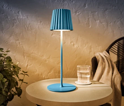 Modrá LED venkovní dobíjecí stolní lampa stojí na stole vedle sklenice vody.