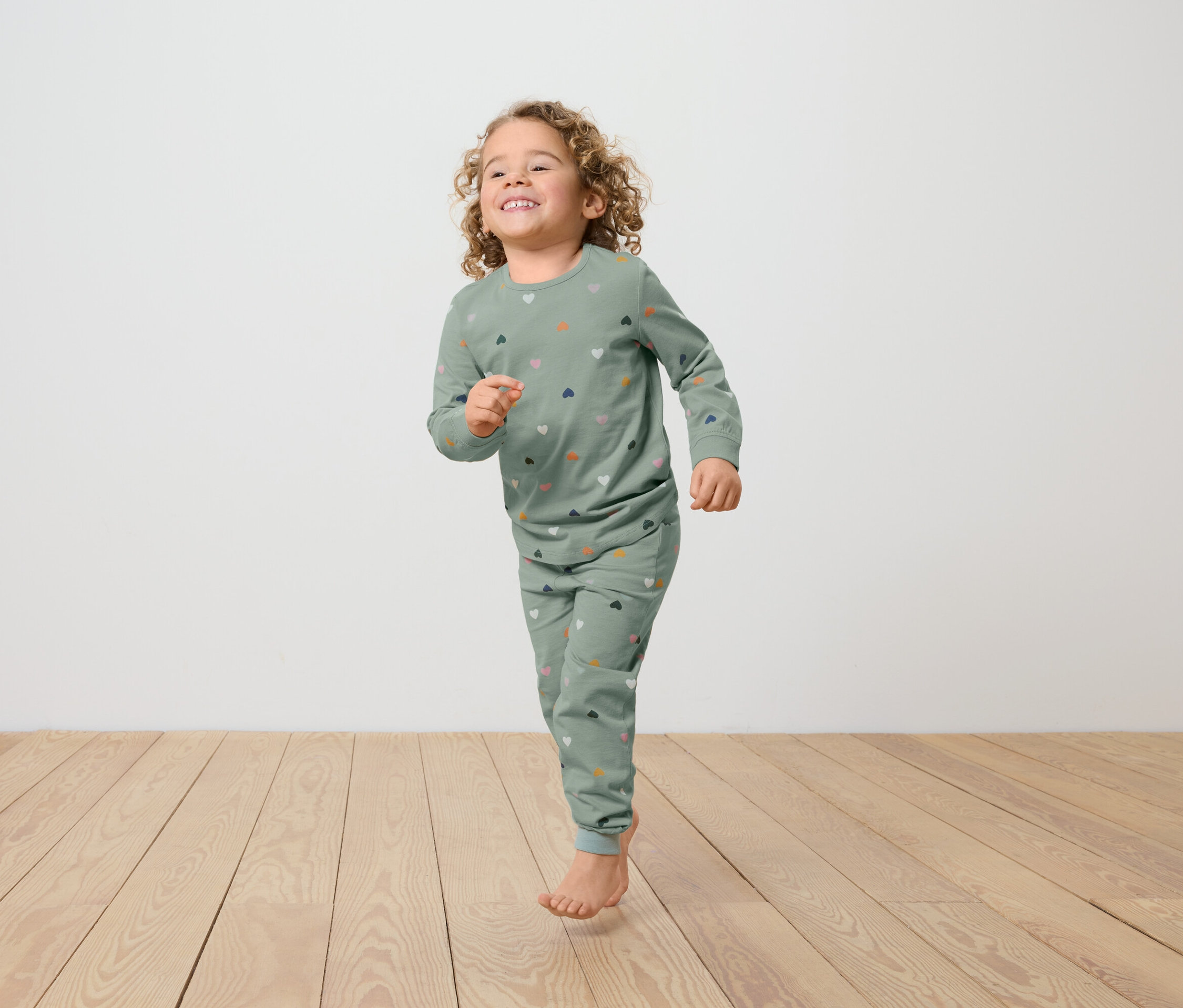 Dívka s kudrnatými vlasy v zeleném pyžamu s potiskem srdcí běží po dřevěné podlaze.