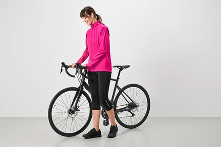 Žena v růžové bundě, černých cyklistických kalhotách a černých botách stojí vedle černého kola.