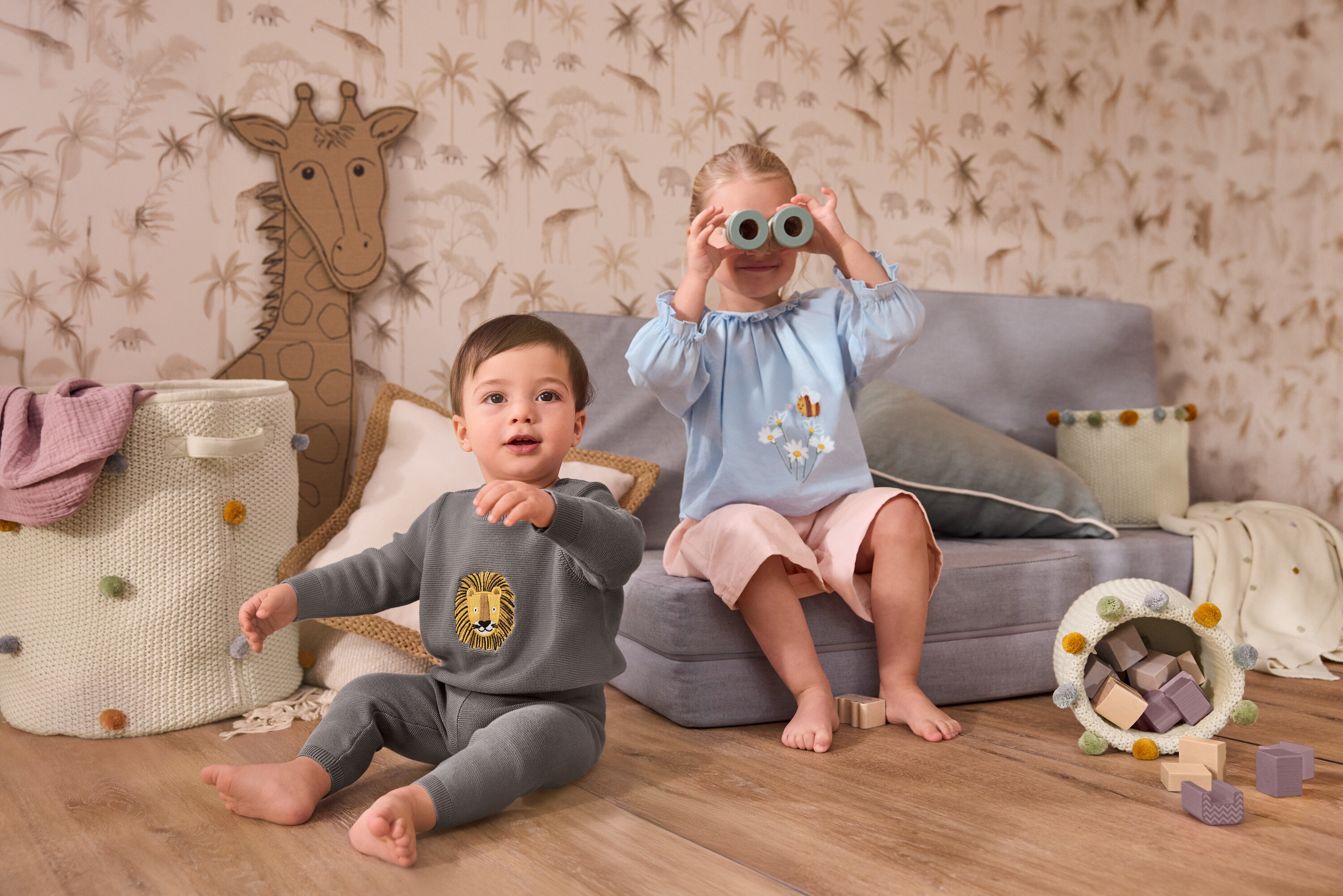 Malé dítě sedí vedle velkého úložného koše do dětského pokoje. Dívka sedí na šedé pohovce a drží před obličejem pletenou deku pro miminka. Má na sobě dívčí halenu z šambré a dětské manšestrové kalhoty culotte.