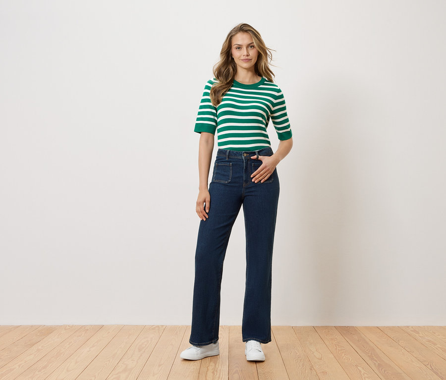 Žena stojí s rukou v kapse kalhot před bílou stěnou. Má na sobě zeleno-bílý pruhovaný top, tmavé džíny a bílé boty.
