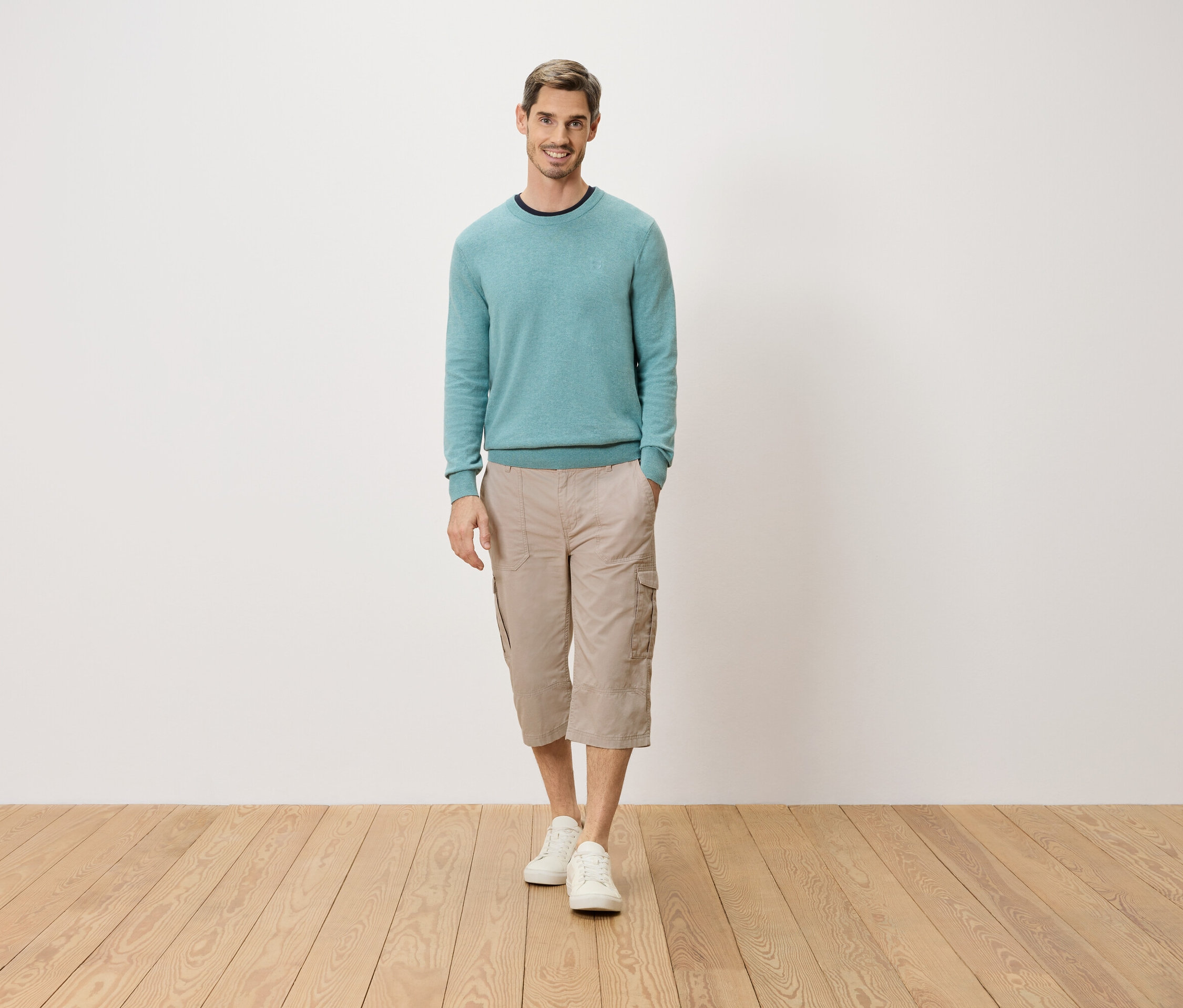 Muž stojí s rukou v kapse před bílou stěnou. Má na sobě světle modrý svetr, béžové tříčtvrťové cargo kalhoty a bílé boty.