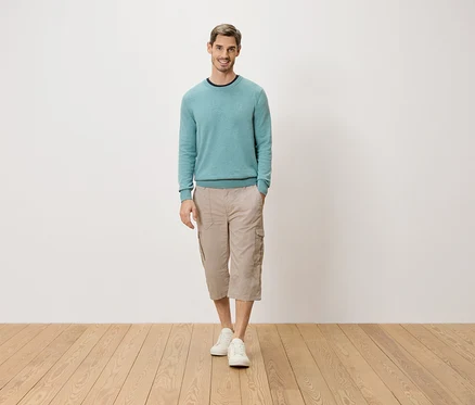 Muž stojí s rukou v kapse před bílou stěnou. Má na sobě světle modrý svetr, béžové tříčtvrťové cargo kalhoty a bílé boty.
