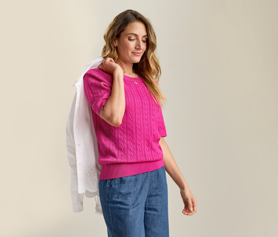 Žena s džínovou bundou s anglickou krajkou přes rameno a růžovým svetrem z jemného úpletu s copánkovým vzorem a džínovými kalhotami.