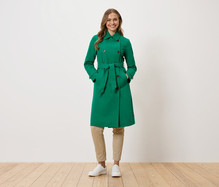 Žena stojí s rukama v kapsách zeleného trenčkotu.